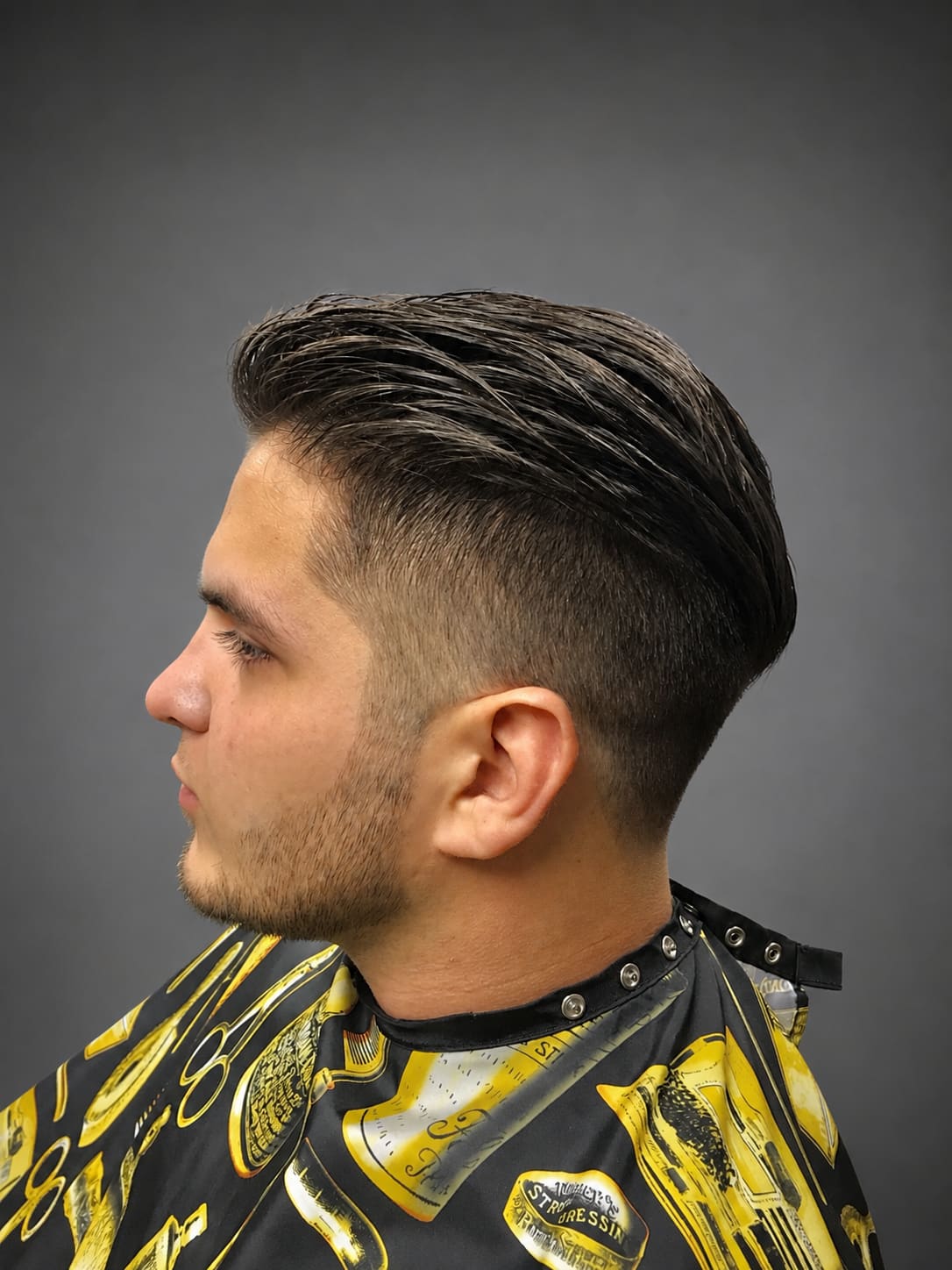 A dark slicked-back cut tapered short above the ear, seen from the side, over a patterned barber cape.